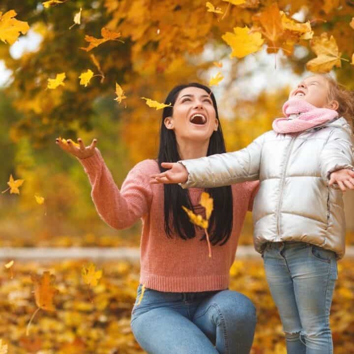 An adult and a child stand outdoors under a tree, joyfully tossing autumn leaves in the air. Both are smiling and dressed in warm clothing.
