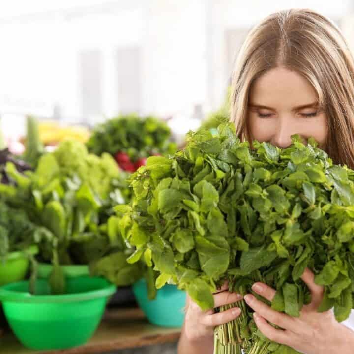 A woman stands in a market holding and smelling a large bunch of fresh green herbs, with other vegetables and greens visible in the background.
