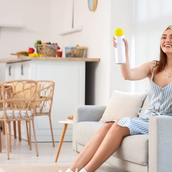 Woman sitting on a couch in a bright kitchen holding up a water bottle and smiling, with a table and chairs visible in the background.