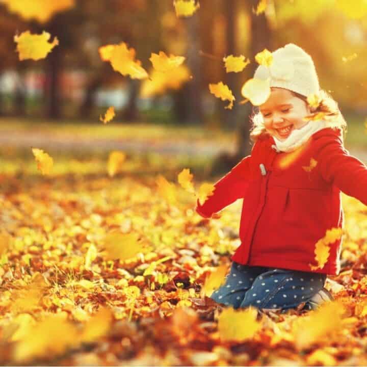 A child in a red coat and white hat plays among falling autumn leaves in a sunlit park.