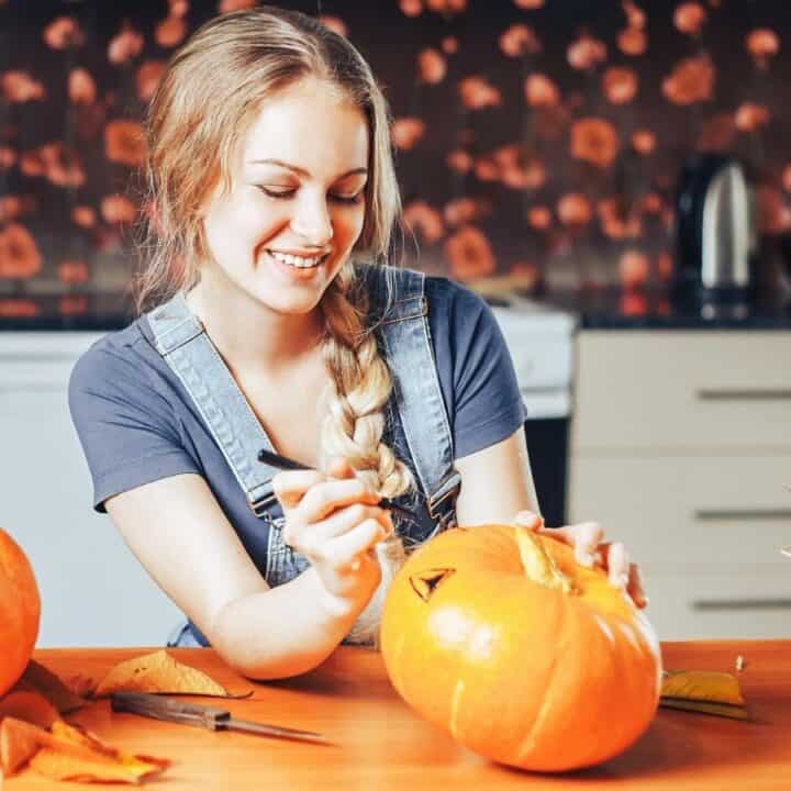 A woman sits at a kitchen table carving a face into a pumpkin, surrounded by other pumpkins, autumn leaves, and a wicker basket.