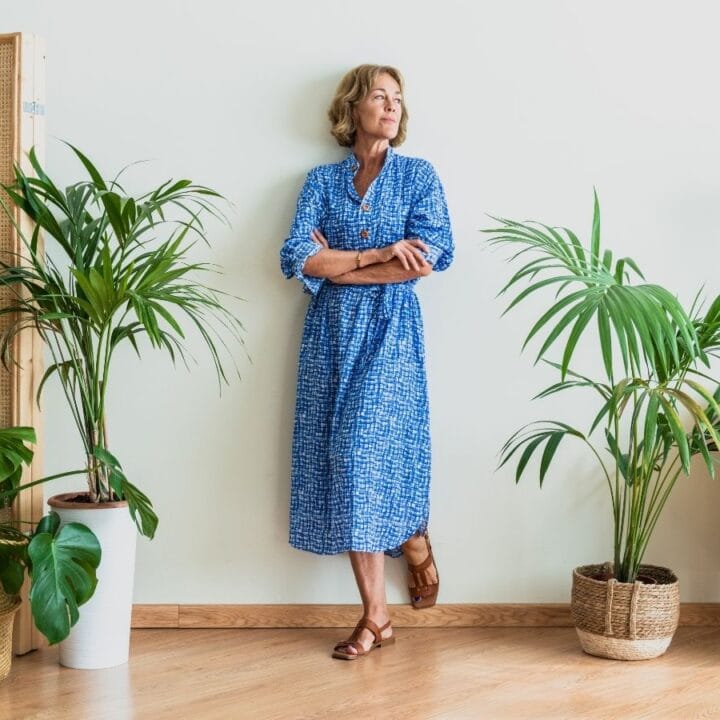 A woman in a blue dress stands with arms crossed, leaning against a white wall, surrounded by potted plants, a wooden screen, and a side table with a lamp.