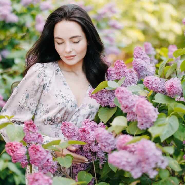 A woman with dark hair in a light floral dress stands among blooming lilac bushes, surrounded by vibrant purple flowers and green leaves.