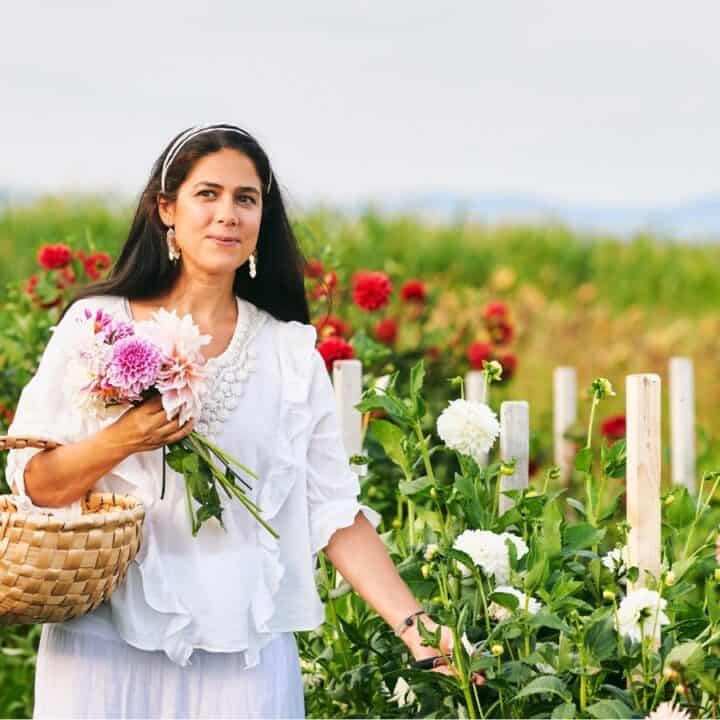 A woman in a white outfit holds a basket and a bouquet of flowers while standing in a flower field with blooming plants and greenery in the background.