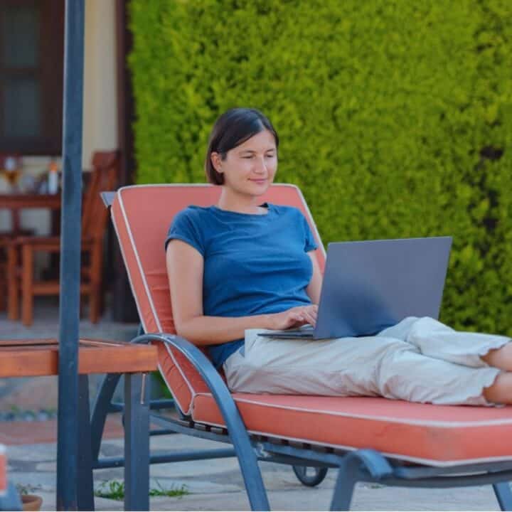 A person sits on a lounge chair outdoors with a laptop on their lap, working or browsing. There are green bushes and patio furniture in the background.