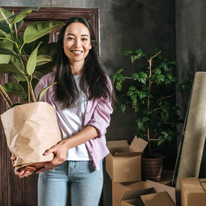 A woman stands in a room with moving boxes, holding a potted plant and smiling at the camera.