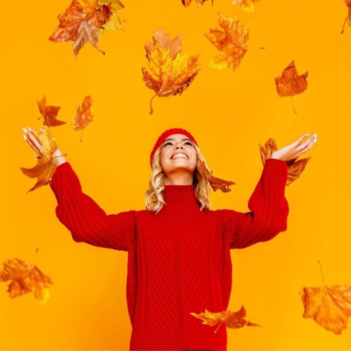 A woman in a red sweater and hat stands smiling with arms raised as autumn leaves fall around her against an orange background.