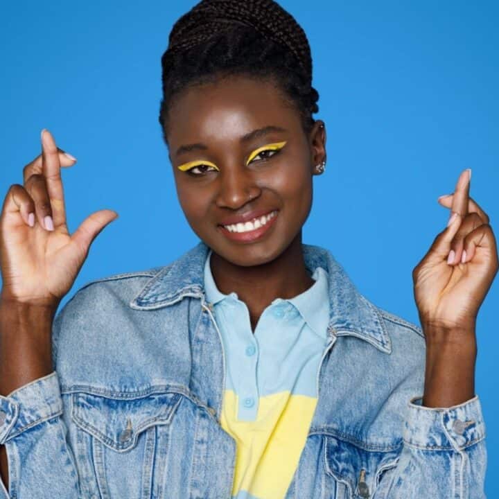 A woman in a denim jacket stands against a blue background, smiling and holding both hands up with fingers crossed.