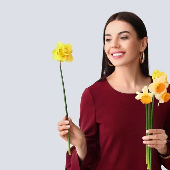 A woman in a maroon top holding yellow daffodil flowers and smiling, standing against a light gray background.