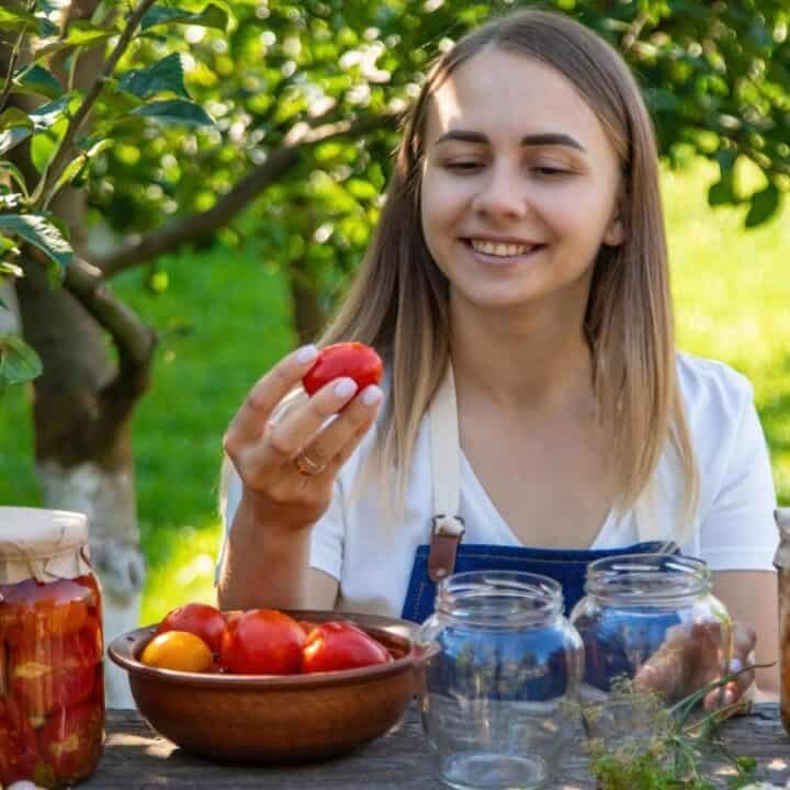 A woman sits outdoors at a table with jars of preserved vegetables, holding a tomato and smiling, surrounded by fresh produce and greenery.