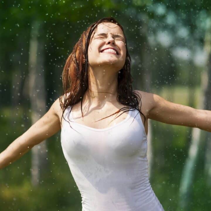 A woman wearing a wet white tank top stands outdoors with arms outstretched, eyes closed, and smiling as rain falls around her in a green, forested area.