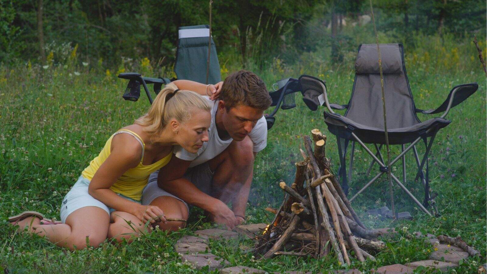 Two people kneel on grass, blowing on a small campfire surrounded by stones, with two folding chairs and trees in the background.
