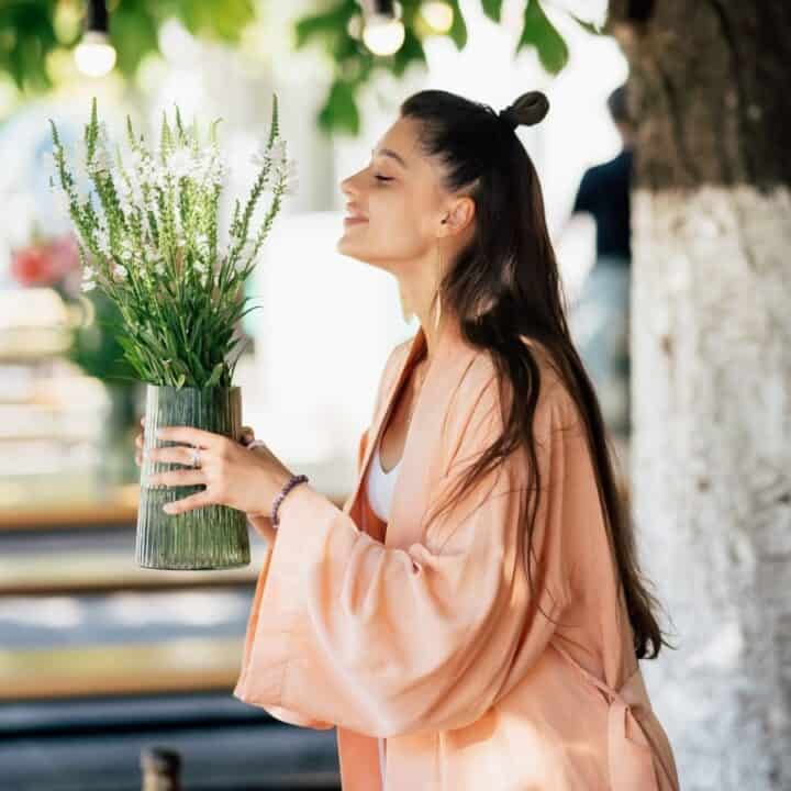 A woman in a light pink robe holds a vase of flowers outdoors, standing near a tree and wooden benches.