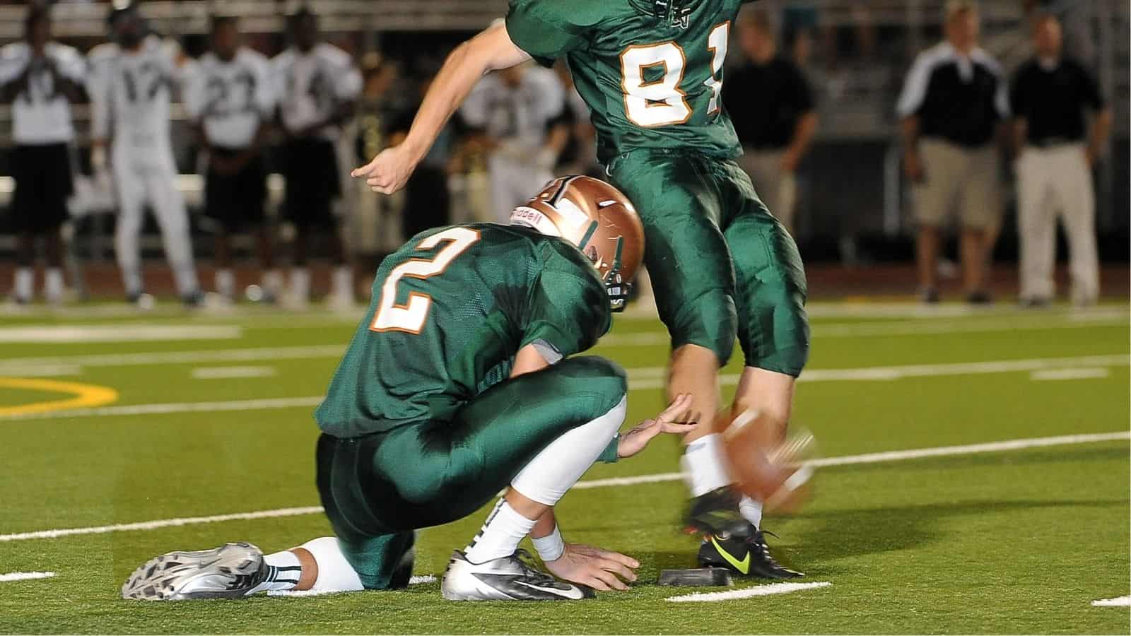 A football player in a green uniform kicks a football held by a teammate during a game on a field, with coaches and players in the background.