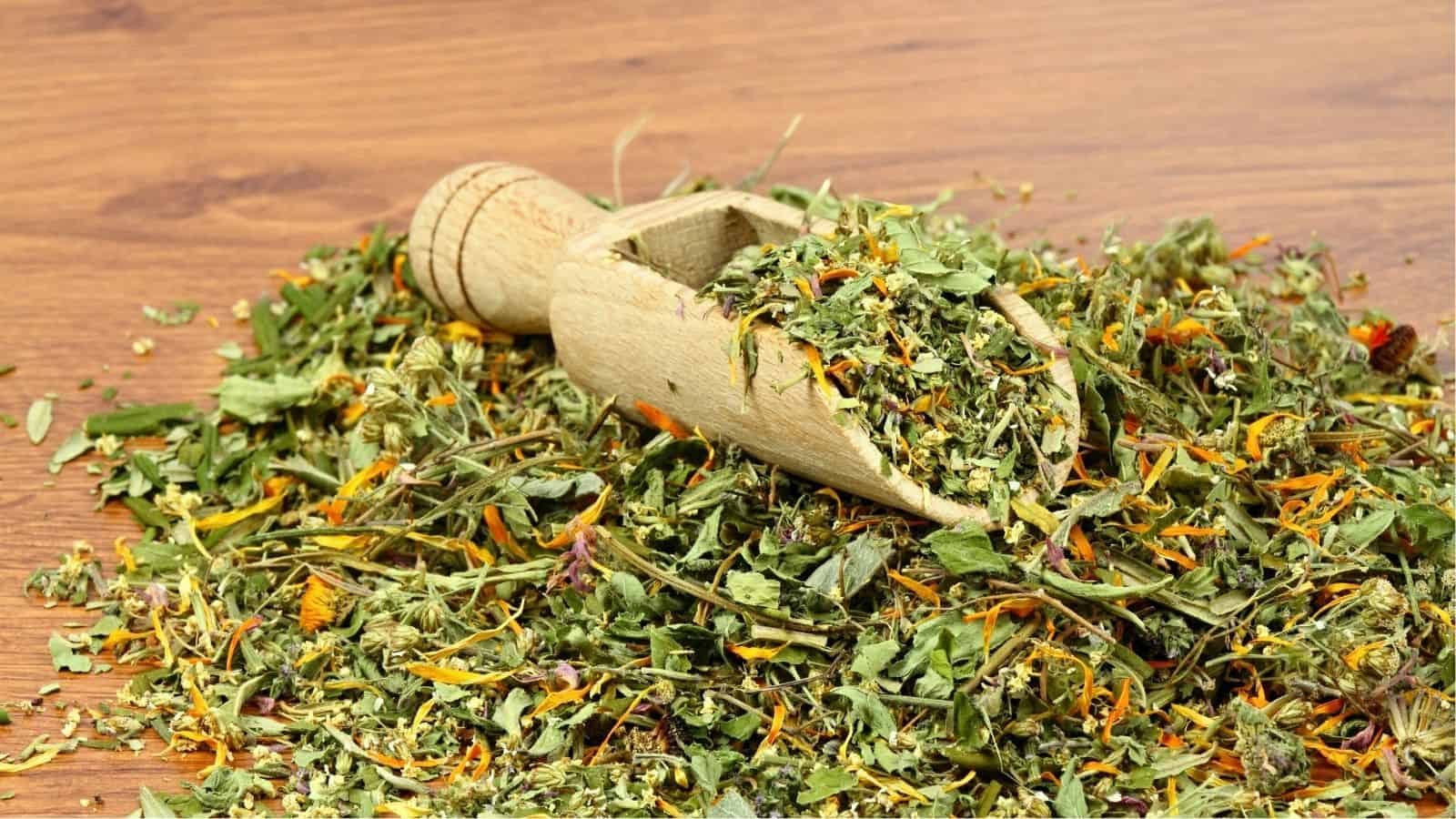 A wooden scoop resting on a pile of dried mixed herbs and flowers on a wooden surface.