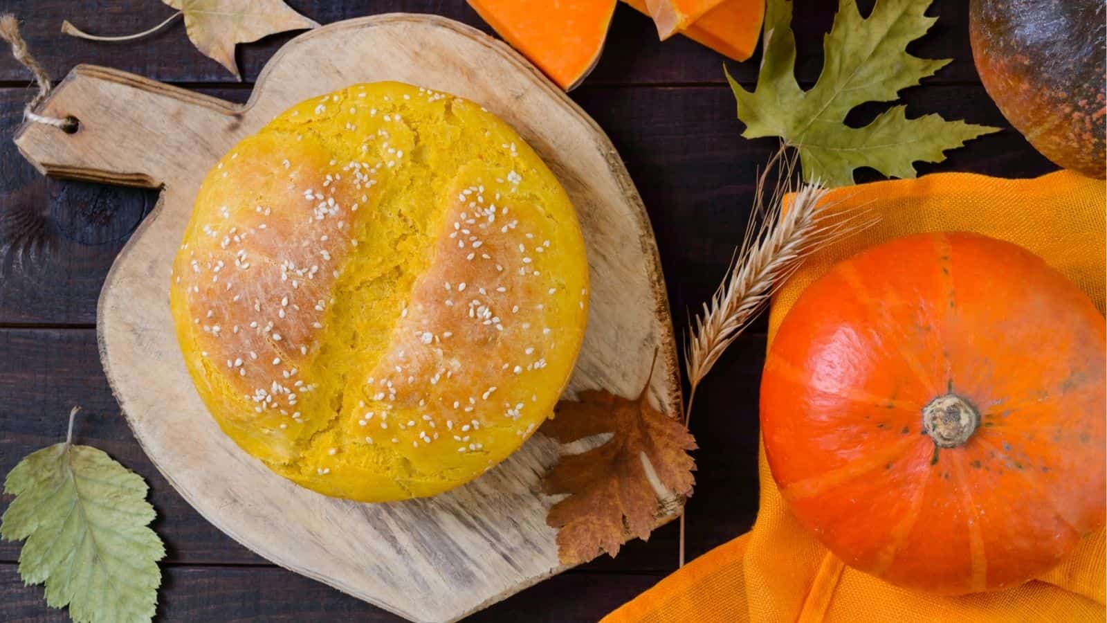 Round loaf of pumpkin bread with sesame seeds on a wooden board, surrounded by pumpkins, leaves, and autumn decorations.