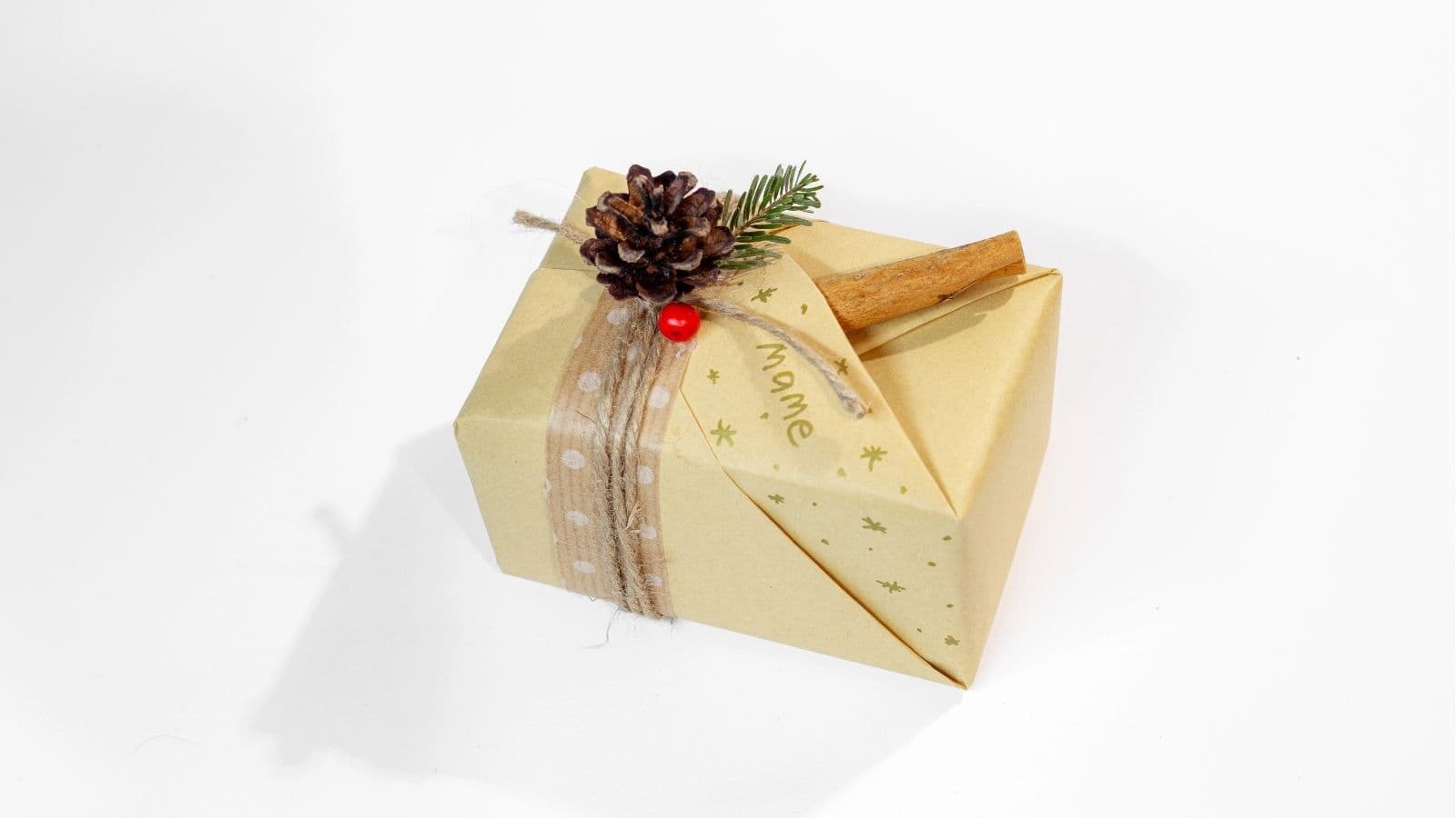 A gift wrapped in beige paper with a polka dot ribbon, decorated with a pinecone, cinnamon stick, greenery, and a red berry, labeled "name" on top.