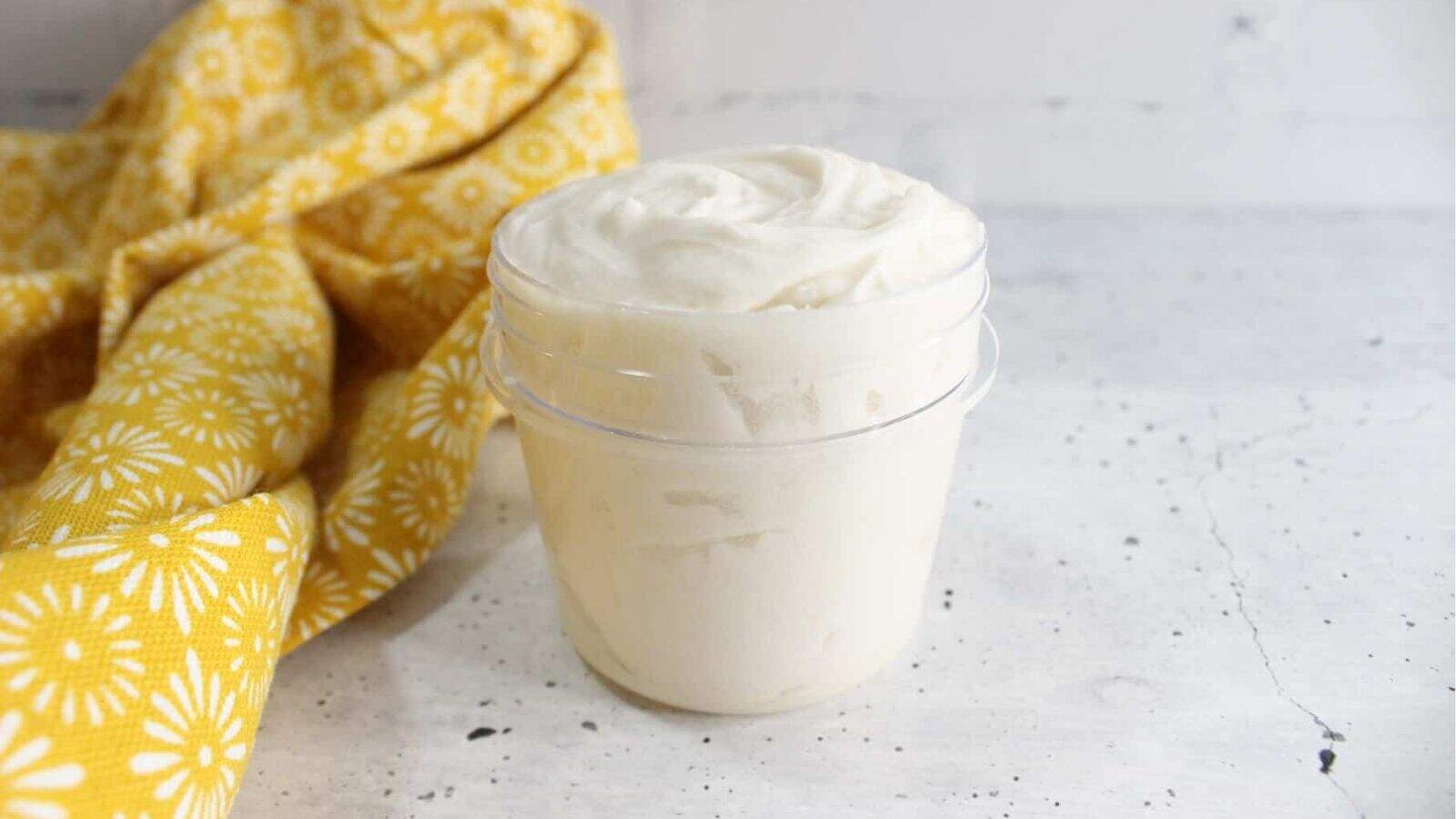 A clear plastic container filled with whipped white cream sits on a light surface next to a yellow patterned cloth.