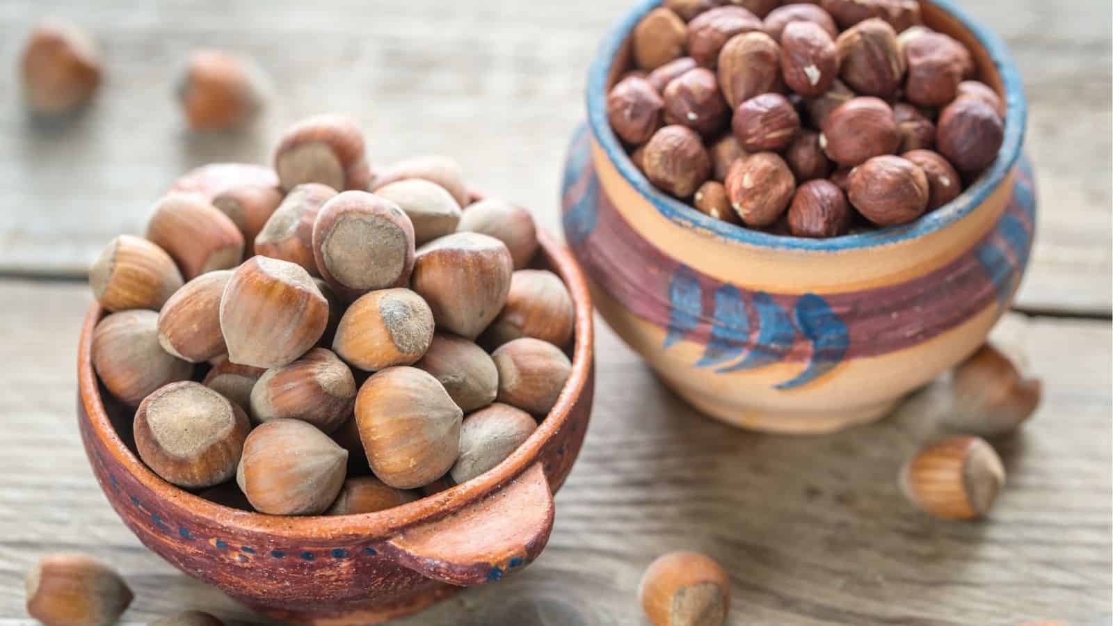Two ceramic bowls filled with hazelnuts, one with shelled nuts and one with unshelled nuts, placed on a wooden surface.