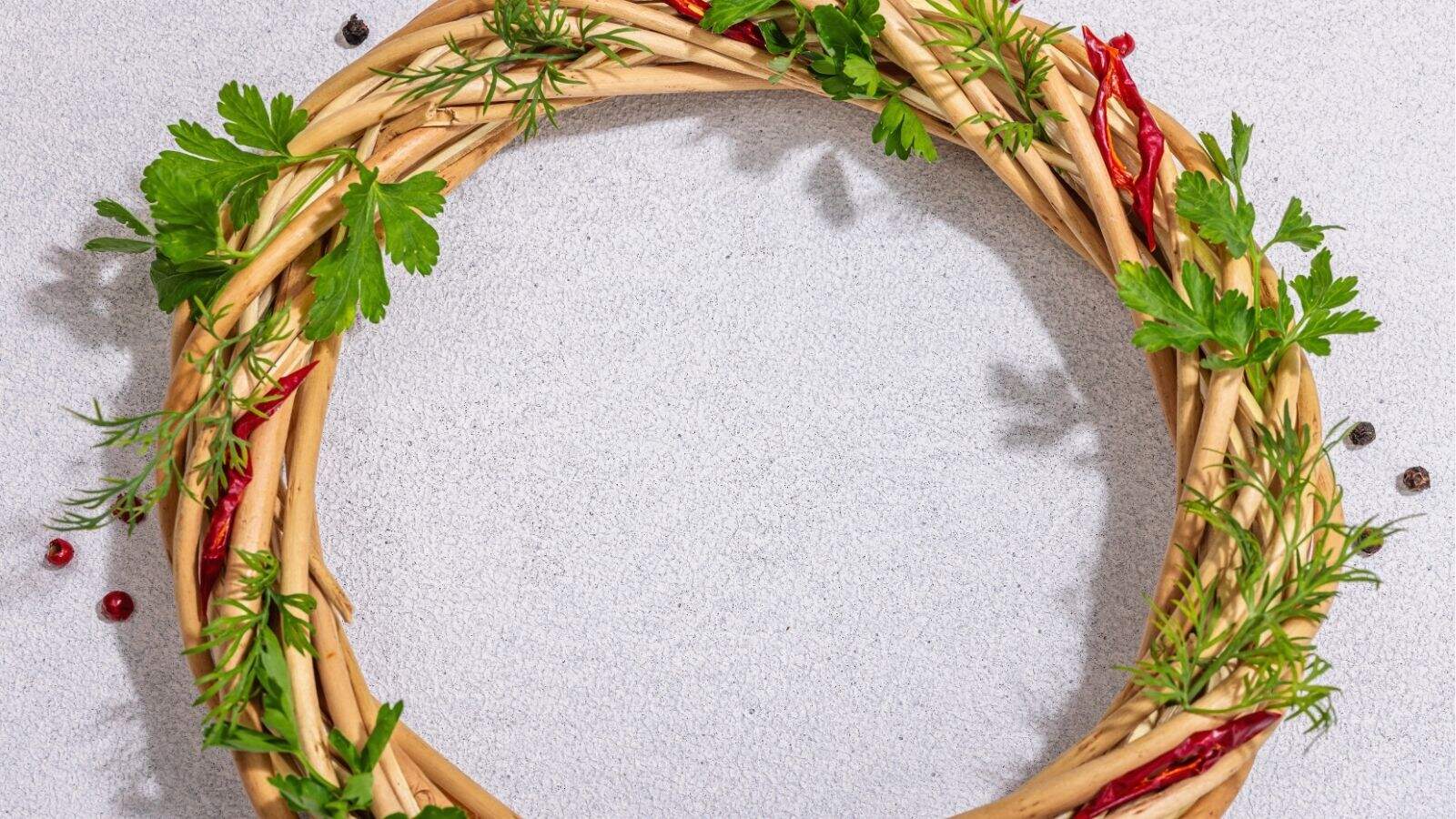 A circular braided wreath made of twigs, decorated with green herbs and red chili peppers, placed on a light gray surface.