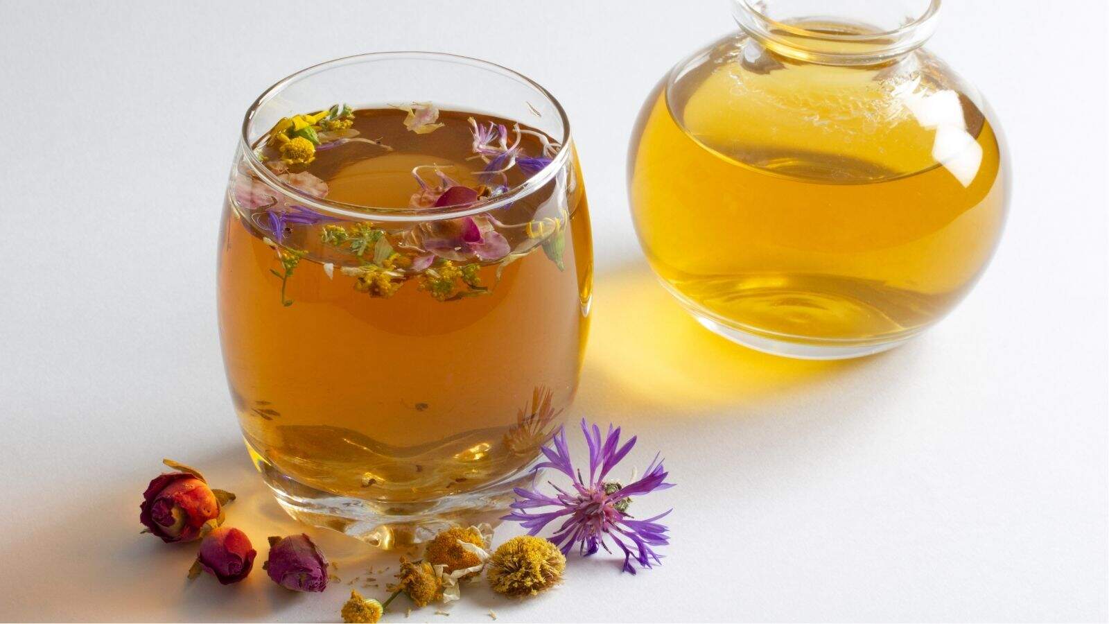 A glass and a small carafe filled with herbal tea, with dried flowers floating in the glass and scattered on the white surface nearby.