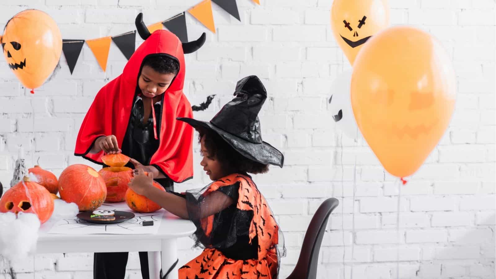 Two children in Halloween costumes, a devil and a witch, sit at a table carving pumpkins. Halloween decorations and balloons are visible in the background.