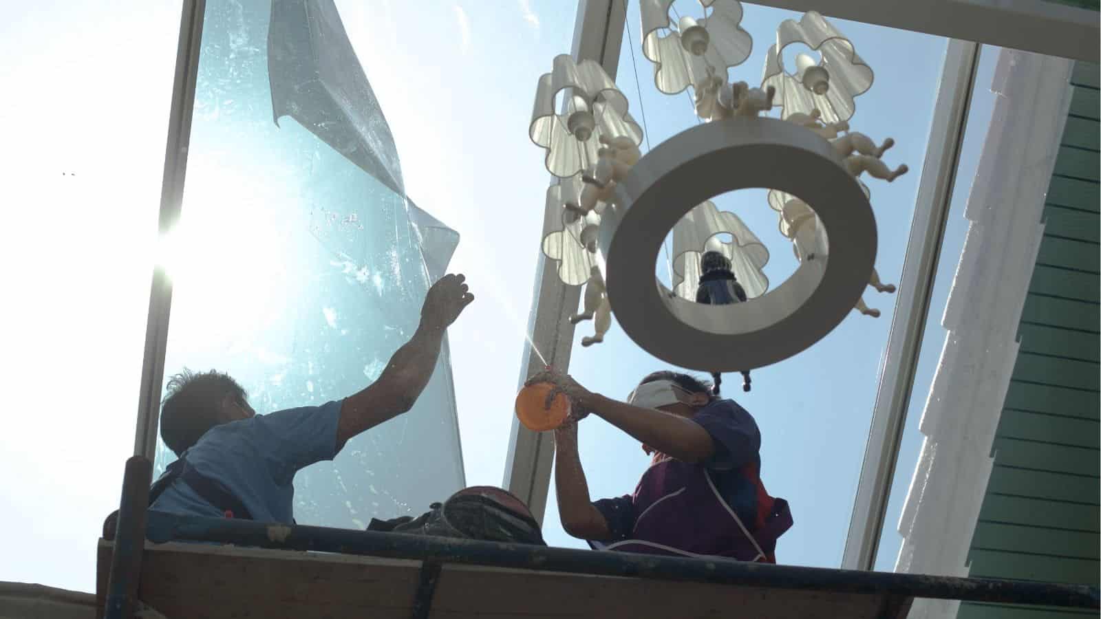 Two workers on a scaffold install or repair a large glass window panel near a ceiling light fixture, with sunlight shining through the glass.