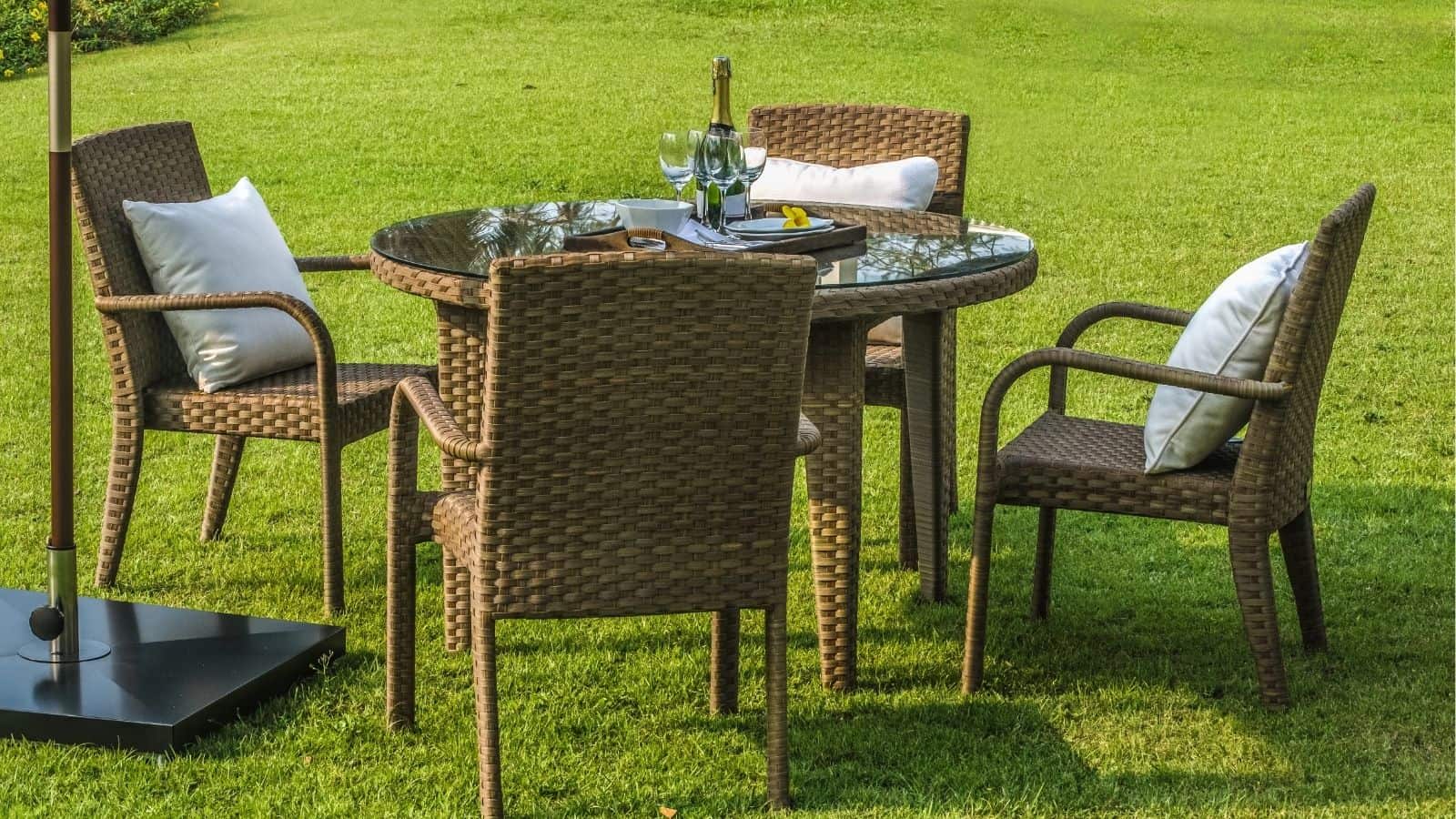 Four wicker chairs with white cushions surround a round glass-top table set with wine glasses and a bottle on a grassy lawn.