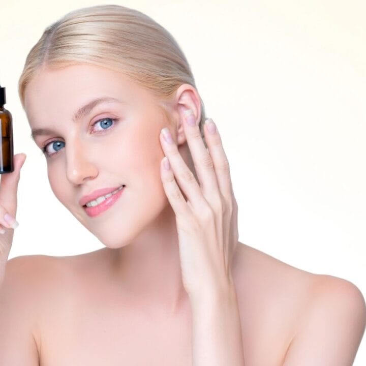 A woman with blonde hair holds a brown dropper bottle near her face and touches her cheek, standing against a light background.