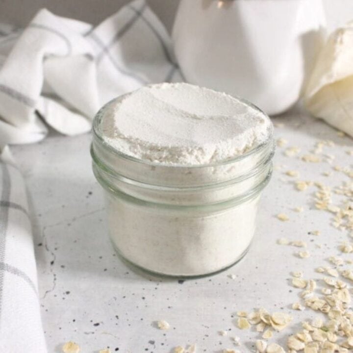 A small glass jar filled with white powder sits on a light surface, surrounded by scattered oats, a cloth napkin, and a white ceramic container.