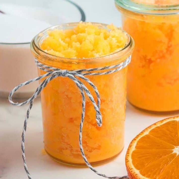 Glass jars filled with orange sugar scrub are displayed on a white surface, surrounded by sliced oranges and a bowl of sugar.