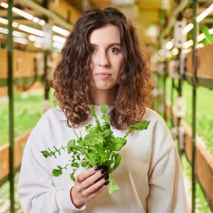 A woman stands in a greenhouse, holding a small bunch of leafy greens, with rows of plants growing on shelves behind her.