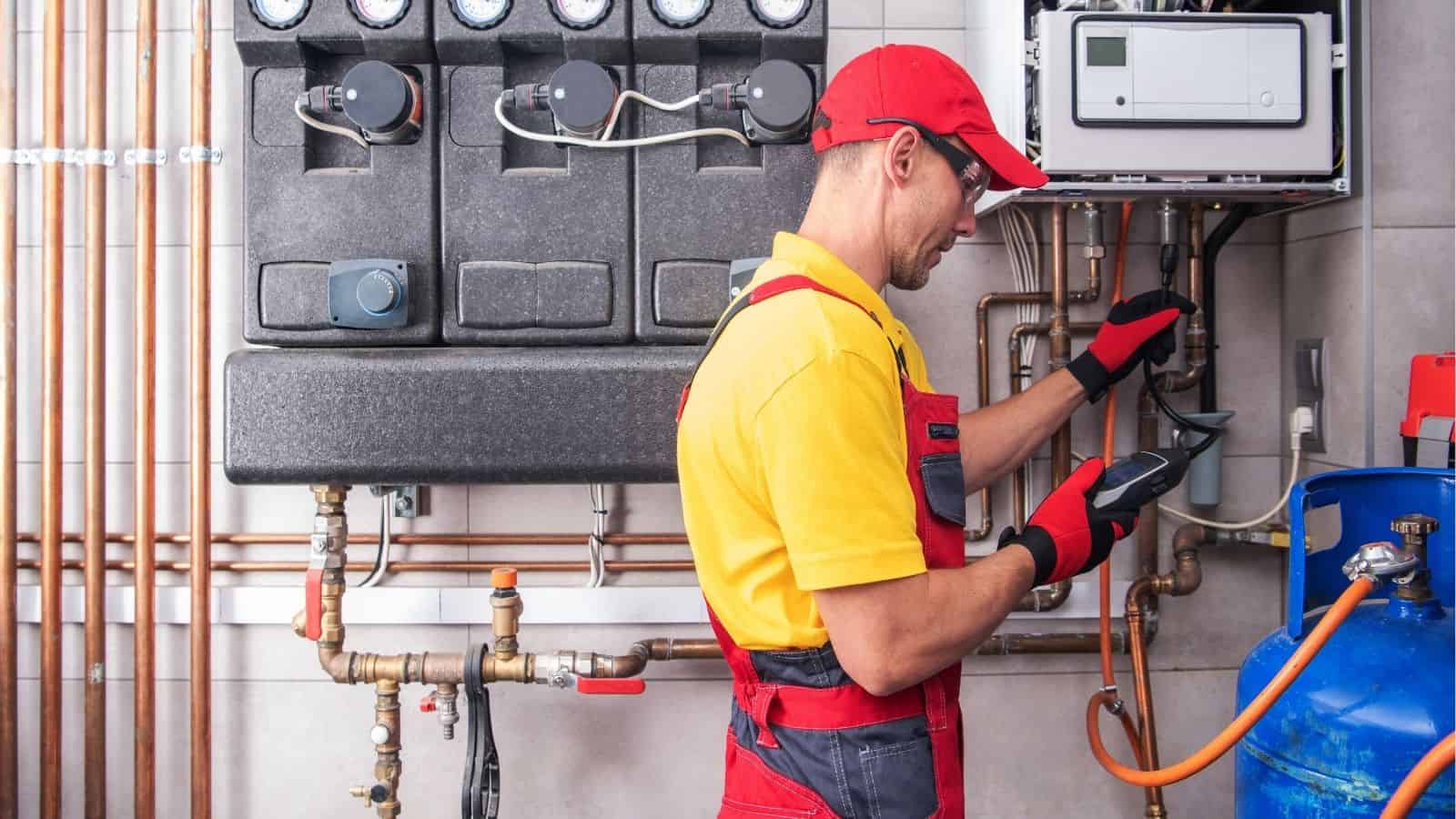 A technician in red overalls and a yellow shirt checks equipment and pipes in a utility room, using a handheld device.