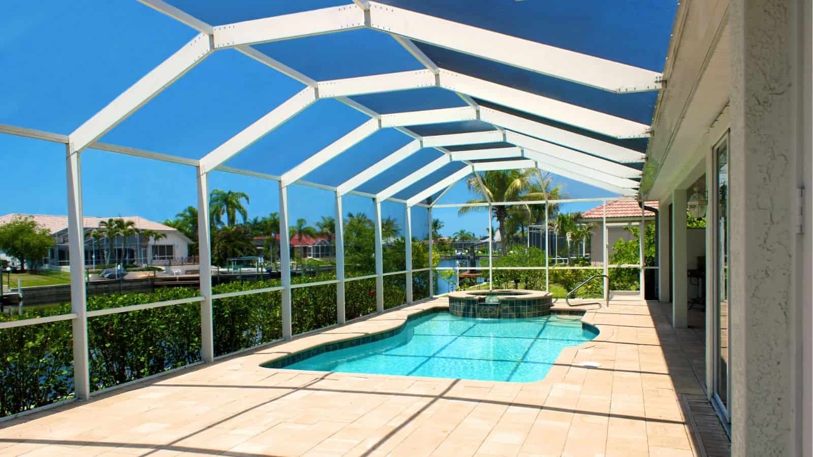 A screened-in patio area with a swimming pool and hot tub, tiled flooring, and views of palm trees and a canal lined with houses under a clear blue sky.