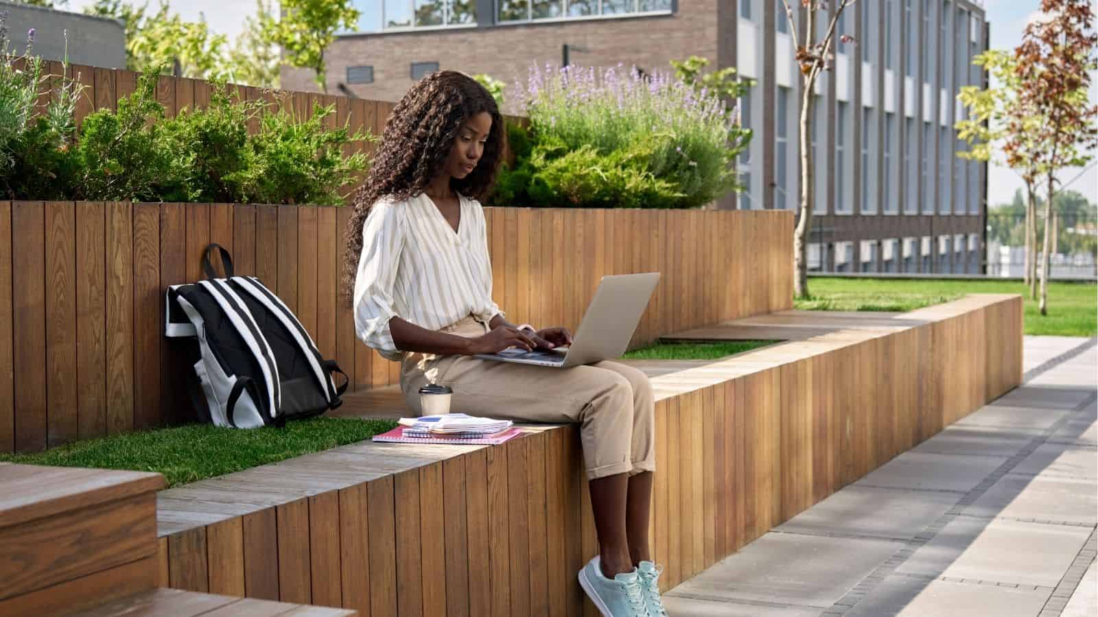 A woman sits on a wooden bench outdoors, working on a laptop with a backpack, books, and a coffee cup beside her.