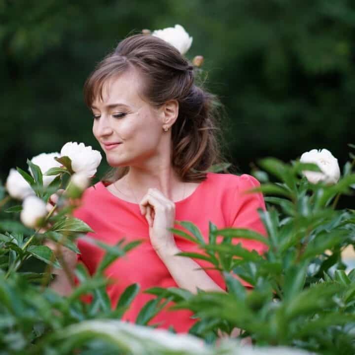 A woman in a pink dress stands in a garden, surrounded by green foliage and white flowers, looking down and smiling gently.