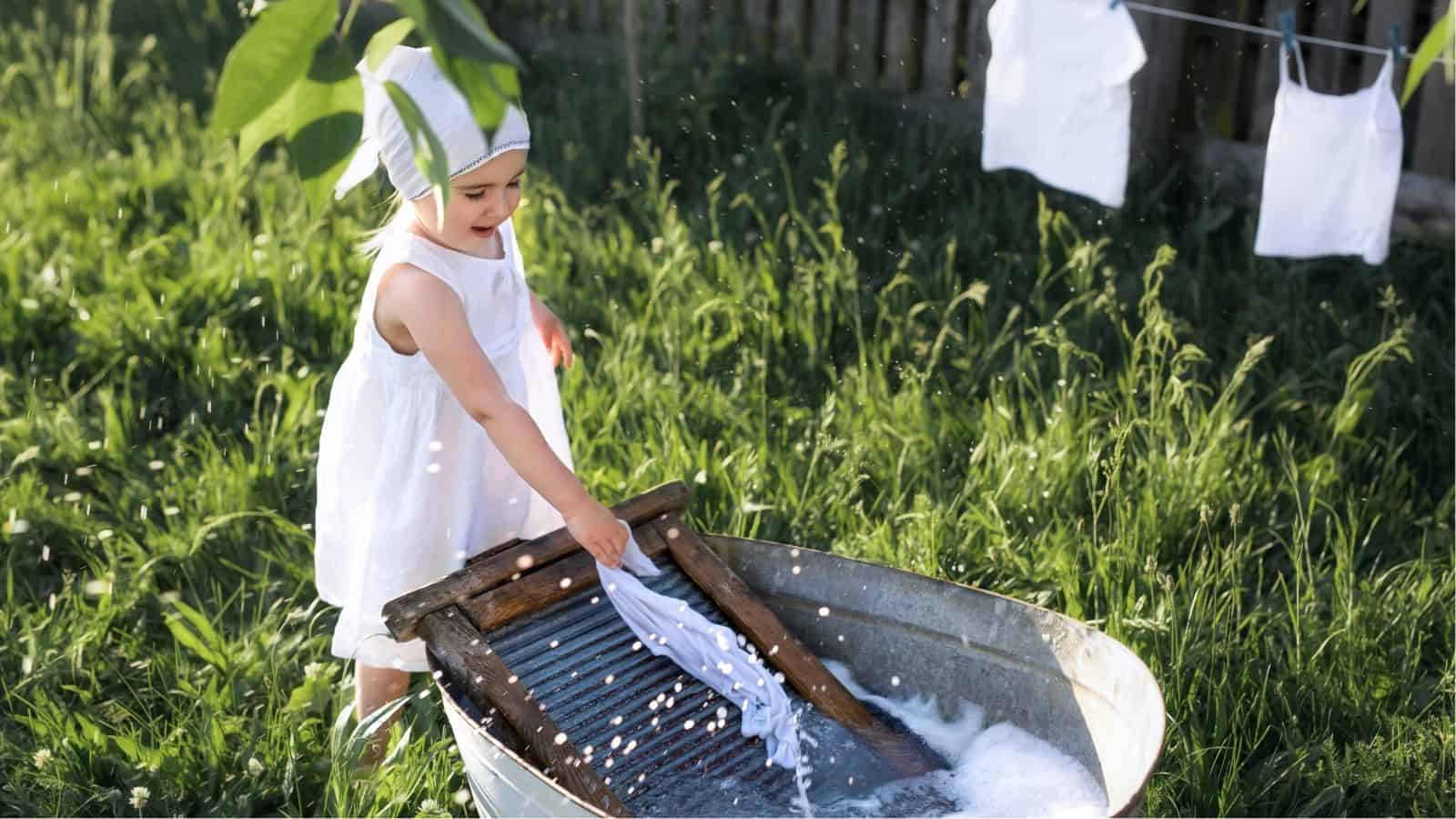 A young child in a white dress and headscarf uses a washboard to clean clothes in a metal tub outdoors, with laundry hanging on a line nearby.