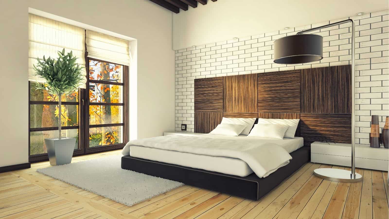 Modern bedroom with wood flooring, a large bed with wood-paneled headboard, floor lamp, potted plant, and large window letting in natural light.