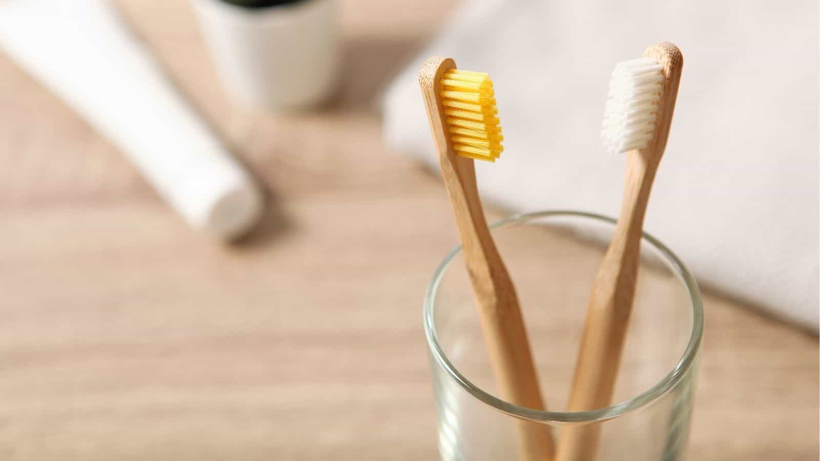Two bamboo toothbrushes with different colored bristles (yellow and white) standing upright in a clear glass on a wooden surface.