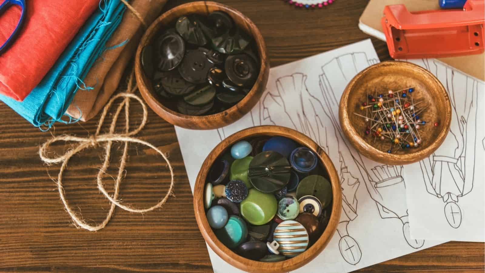 Wooden bowls filled with assorted buttons and sewing pins on a table with fabric, string, scissors, and fashion sketches.