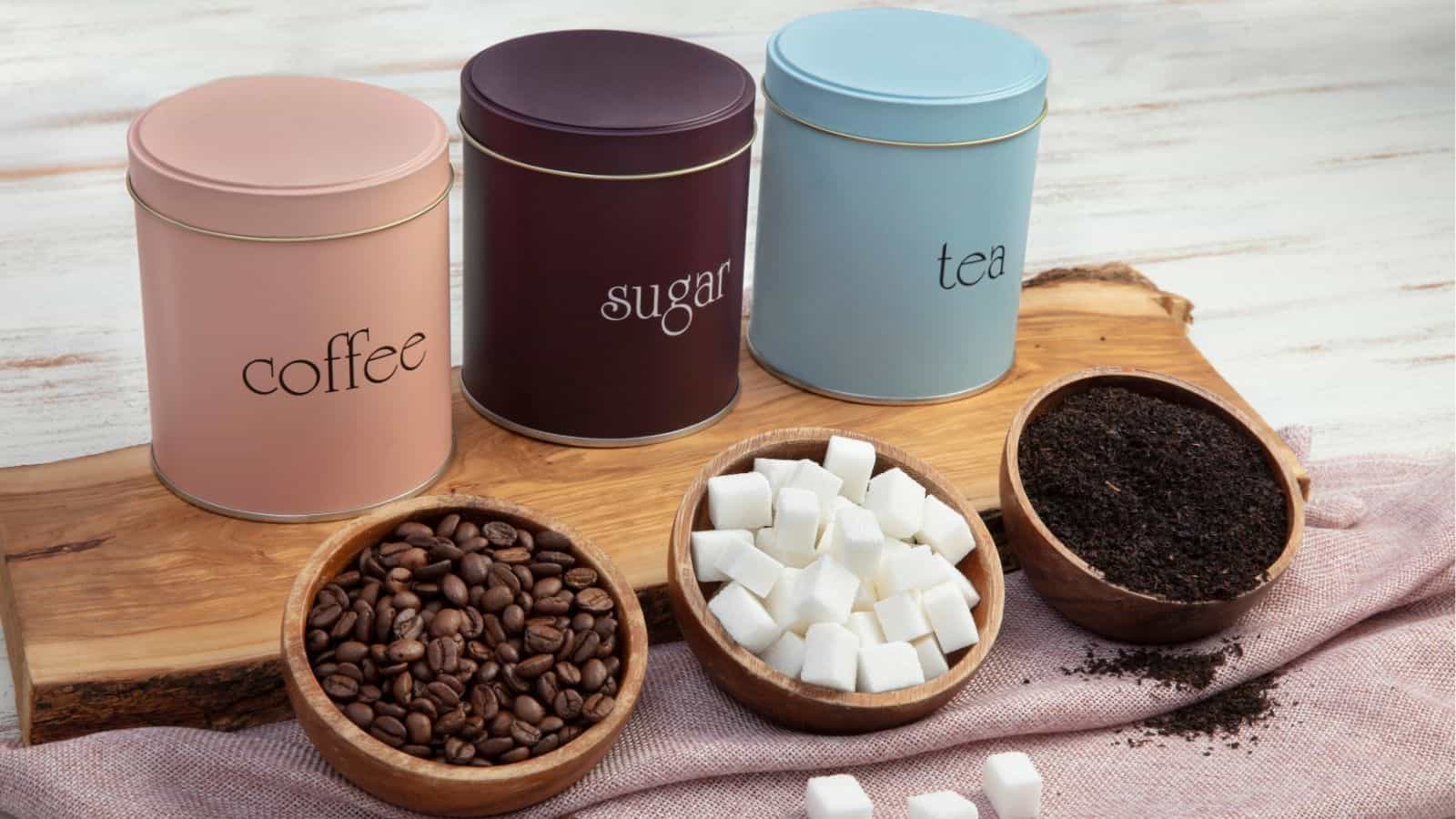 Three labeled canisters for coffee, sugar, and tea are displayed with bowls containing coffee beans, sugar cubes, and loose tea leaves on a wooden board.