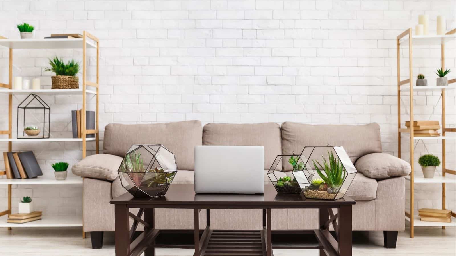 A beige sofa against a white brick wall, flanked by shelves with books and plants. A coffee table with geometric terrariums and an open laptop is in the foreground.