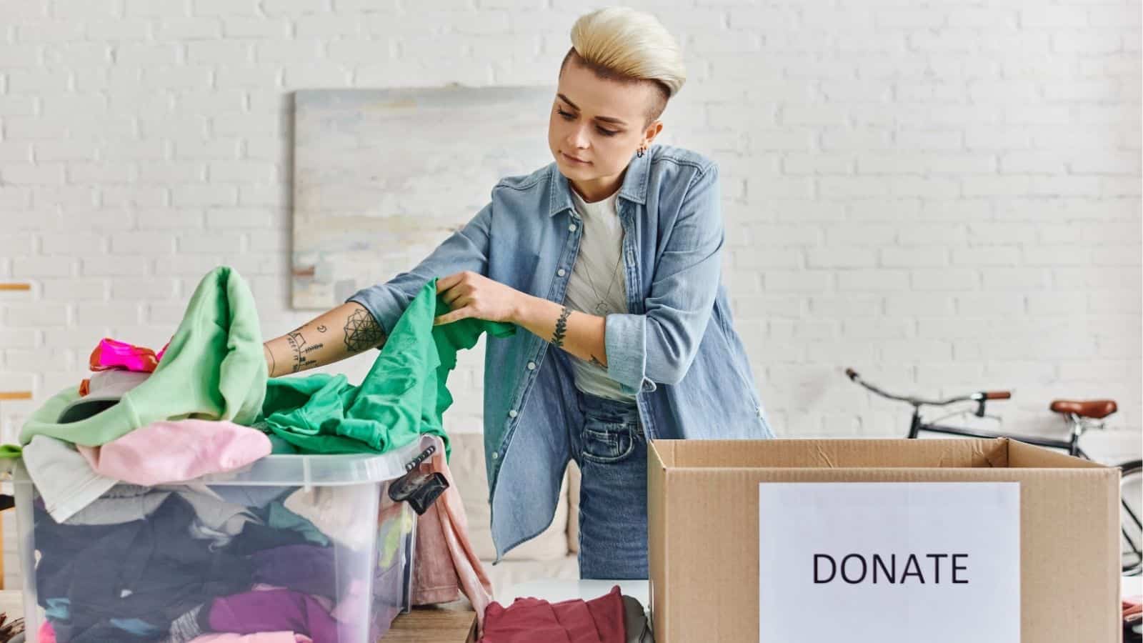 A person sorts through a pile of clothes next to a cardboard box labeled "DONATE" in a bright, indoor setting.