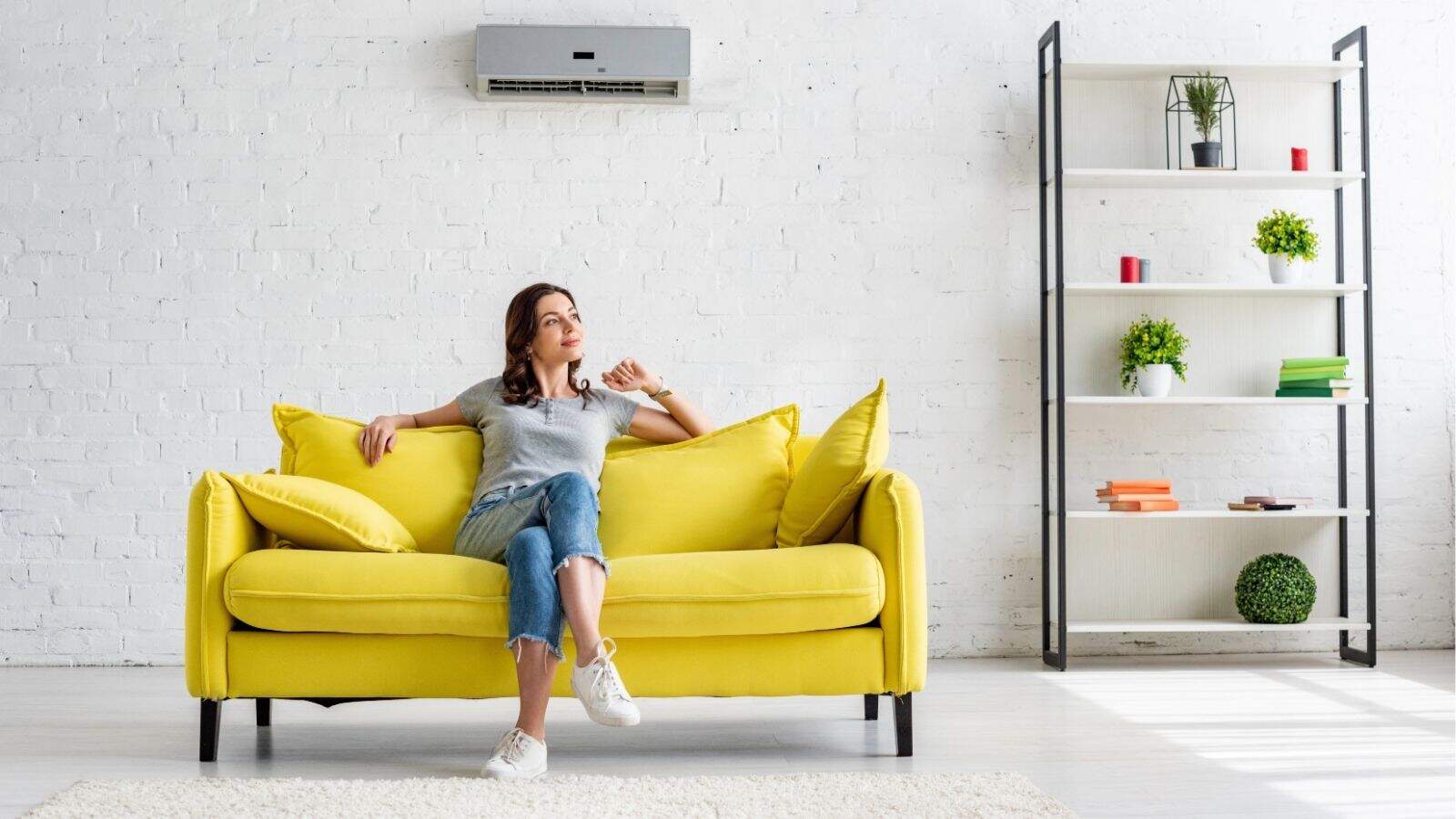 A woman sits on a yellow sofa in a modern living room with a white wall, air conditioner, and a shelving unit holding plants and books.