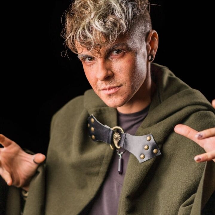 Person wearing a green cloak with a decorative clasp, holding hands up in a dramatic pose against a black background.