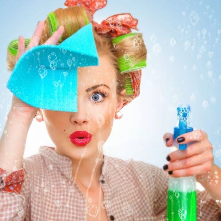 A woman with curlers in her hair cleans a soapy glass surface with a sponge and holds a green spray bottle, looking surprised.