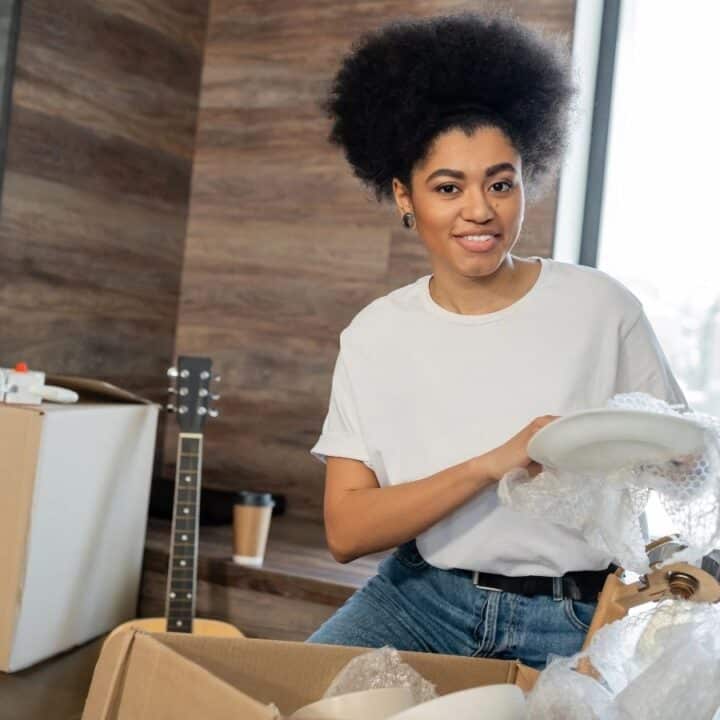 A person in a white shirt packs or unpacks plates with bubble wrap in a room filled with moving boxes, a guitar, and potted plants.