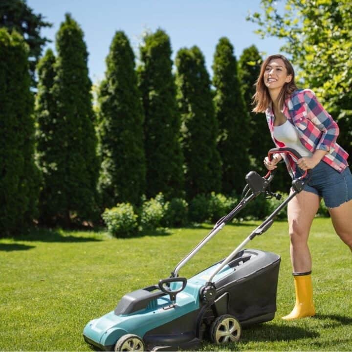 A woman in yellow boots mows the lawn with an electric lawn mower on a sunny day, surrounded by trees and greenery.