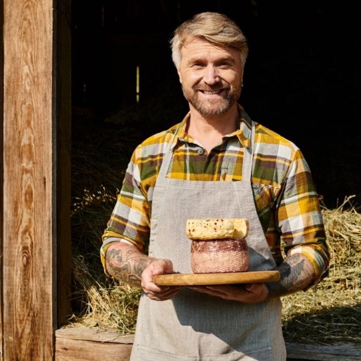 Man in apron standing by a barn door holds a wooden tray with three stacked rounds of cheese, smiling at the camera.