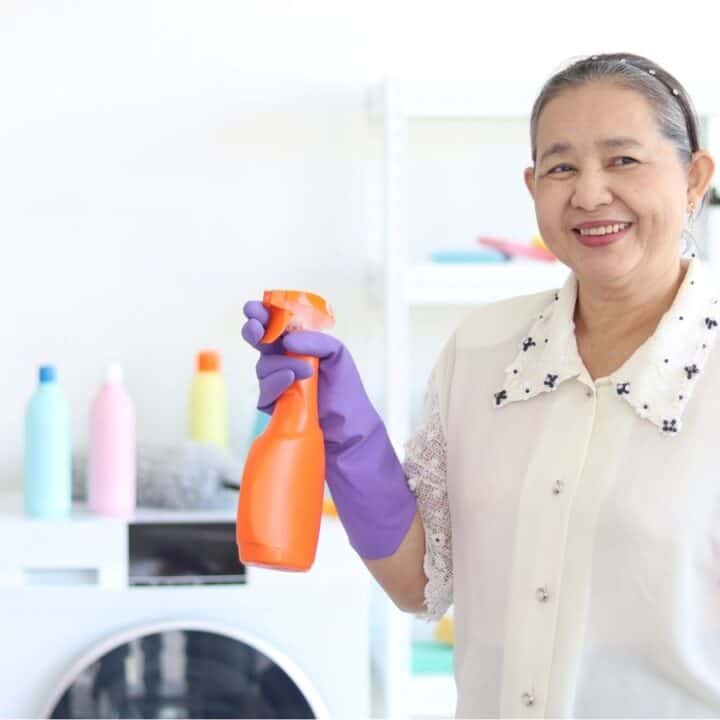 An older woman wearing purple gloves holds an orange spray bottle in a laundry room with shelves, detergent bottles, and folded towels in the background.